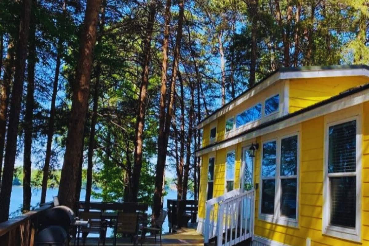 A yellow building with a covered deck, surrounded by tall trees and a body of water.