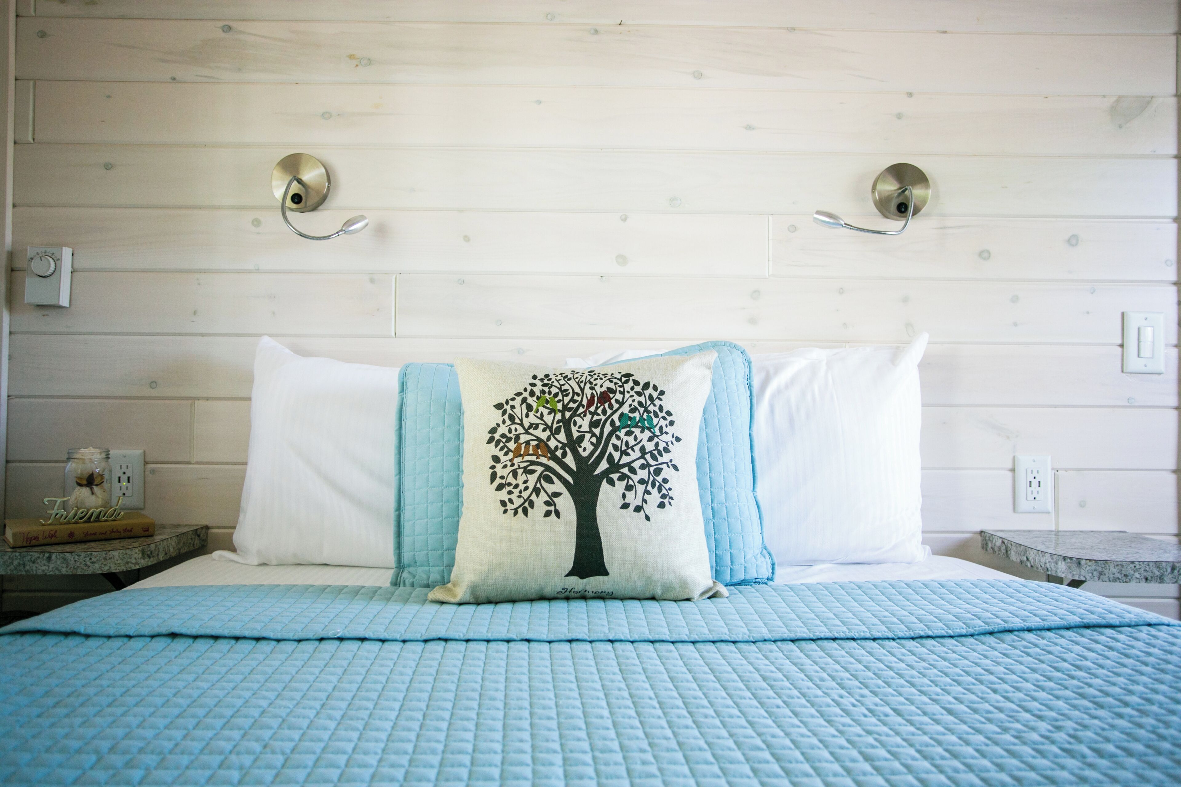 Un dormitorio con cama, un almohadón con diseño de árbol, una mesita de noche y una lámpara fijada a la pared.