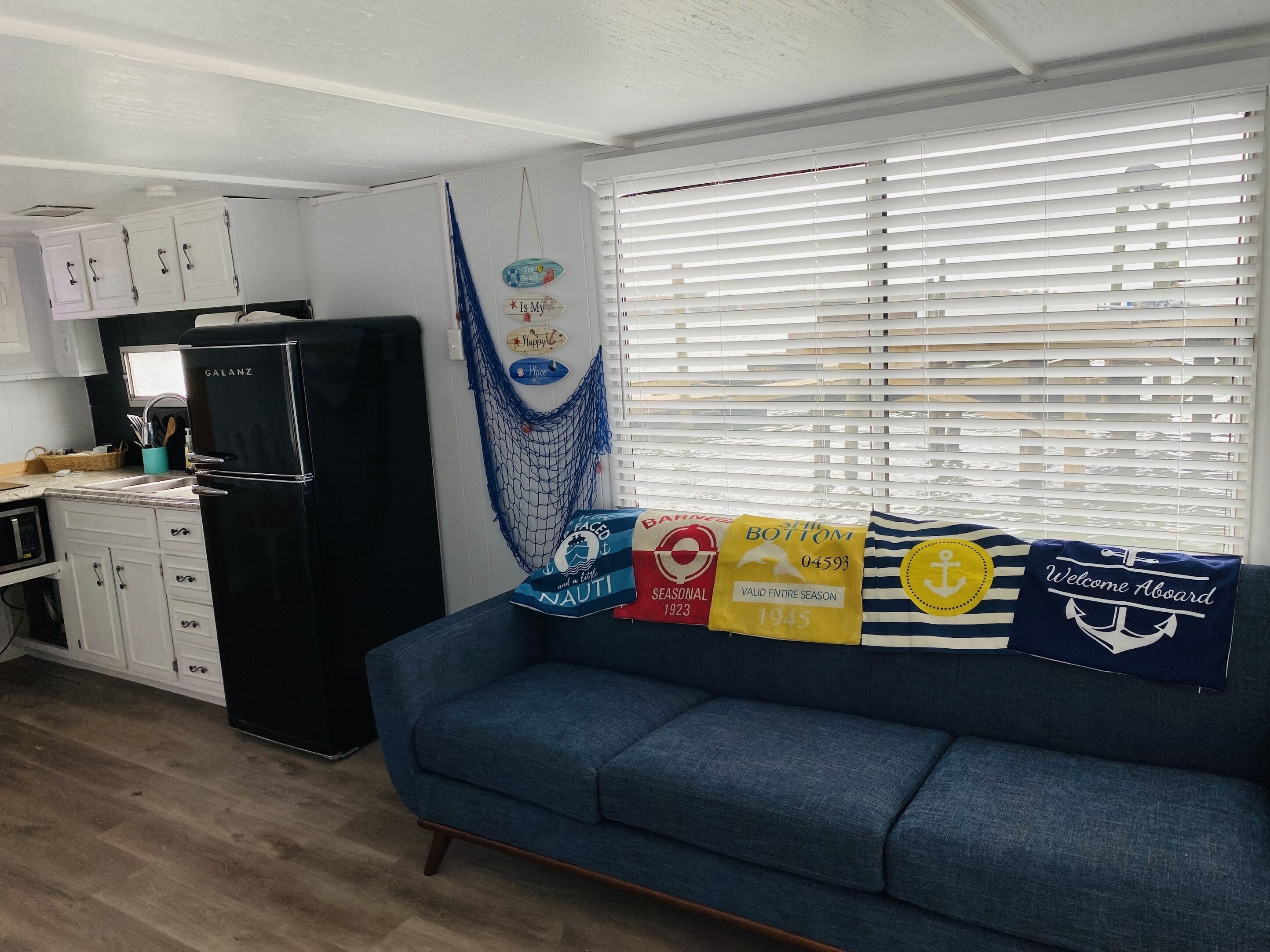 A compact living space with a kitchenette, a blue sofa adorned with nautical-themed cushions, and a hammock.