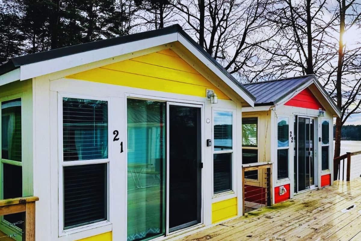 A row of small, colorful cabins with large windows and doors, numbered 21 and 22, on a wooden deck with a view of a lake.