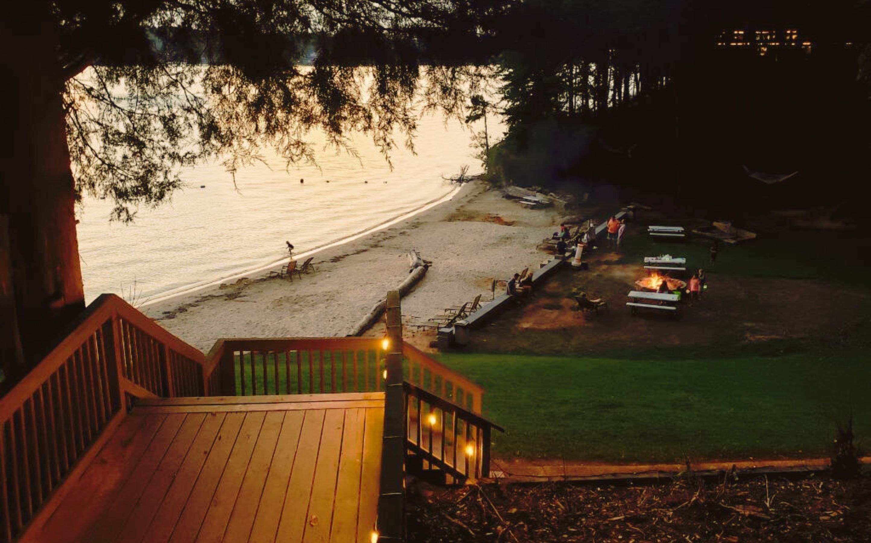 A beach view from a wooden deck overlooking a sandy shore with a dock and a small campfire area.
