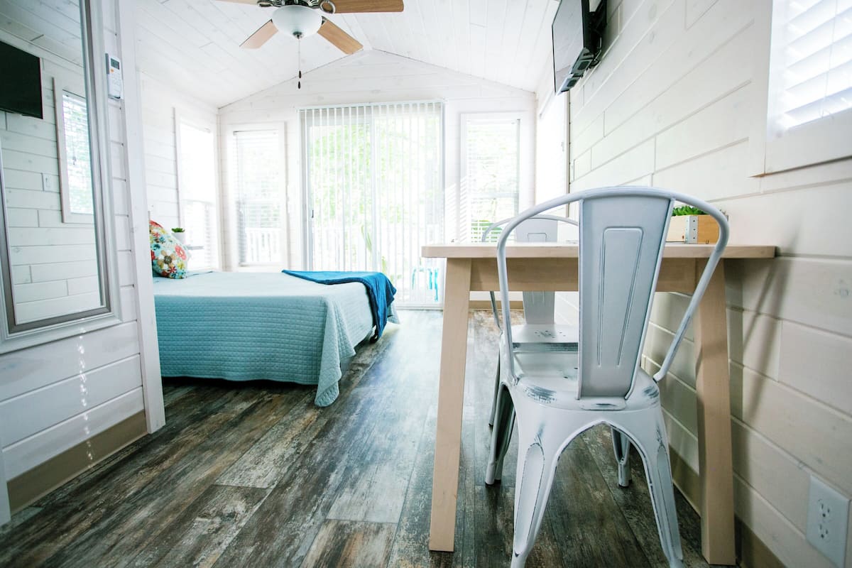 A room with a bed, a desk, and a chair. There is a window with blinds, a ceiling fan, and a door.