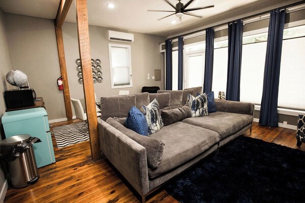 A modern living room with a grey sofa, wooden flooring, a ceiling fan, and a window with curtains.