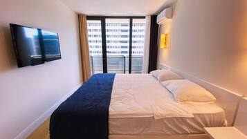 A modern hotel room with a large bed, a flat-screen TV mounted on the wall, and a view of a building through the window.