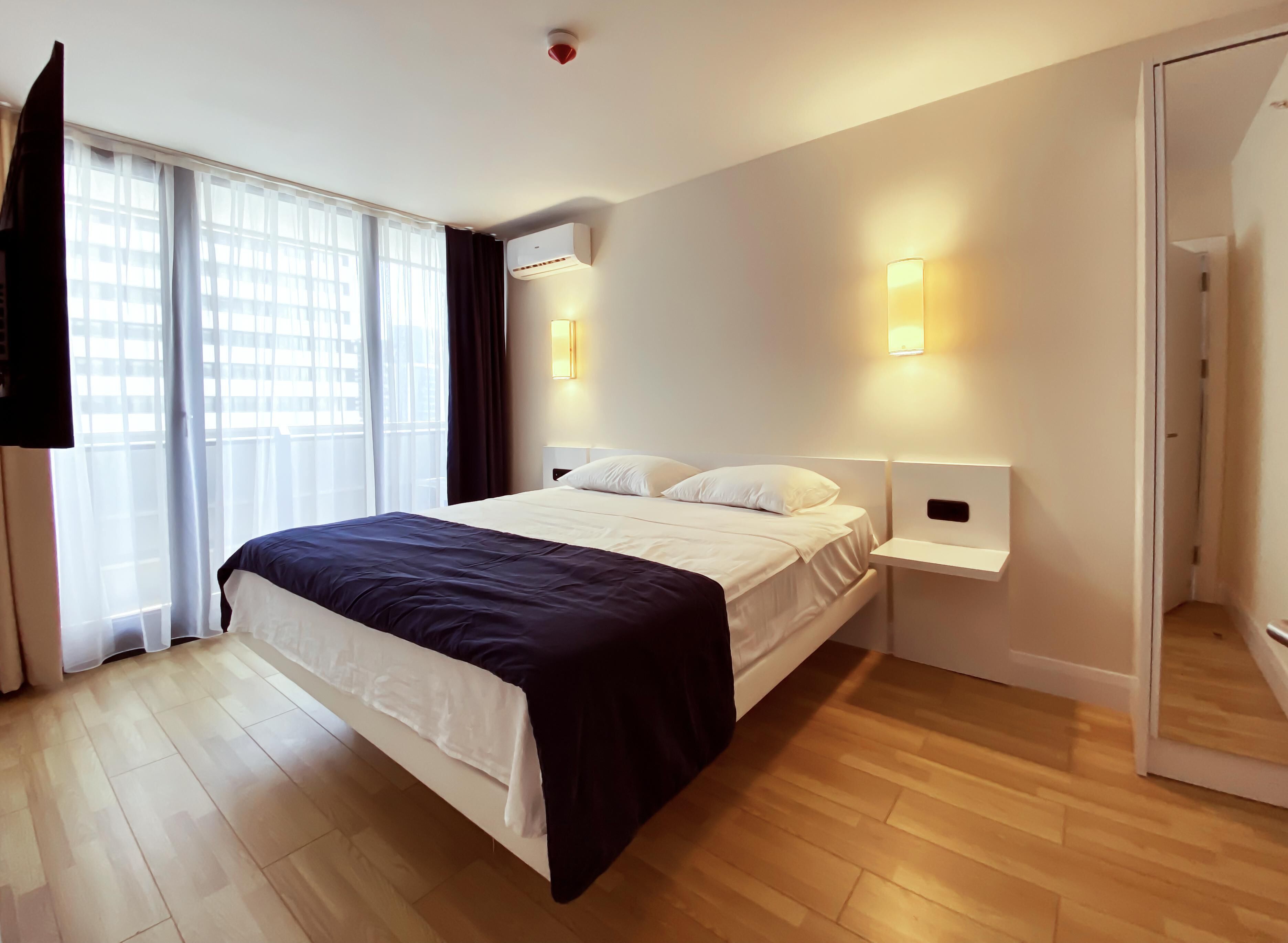 A modern hotel room with a large bed, a flat-screen TV mounted on the wall, a bedside table with a lamp, and a window with sheer curtains.