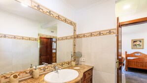 A bathroom with a large mirror, a sink, a vanity, and a wooden door leading to a bedroom. - Juanito (Puerto Pollensa)