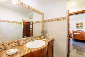 A bathroom with a large mirror, a sink, a vanity, and a wooden door leading to a bedroom. - Juanito (Puerto Pollensa)