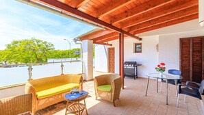 A covered patio area with wicker furniture, a grill, and a view of greenery. - Juanito (Puerto Pollensa)