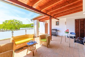 A covered patio area with wicker furniture, a grill, and a view of greenery. - Juanito (Puerto Pollensa)