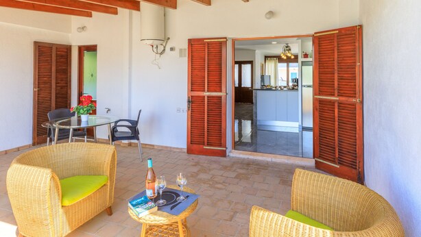 Un patio cubierto con mobiliario de mimbre, una mesa con copas de vino y contraventanas de madera.
