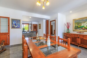 A dining area with a wooden table, chairs, and a glass-top table. There are paintings on the walls and a wooden cabinet with drawers. - Juanito (Puerto Pollensa)