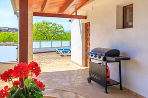 An outdoor patio with a barbecue grill, a view of a body of water, and a balcony with blue chairs. - Juanito (Puerto Pollensa)