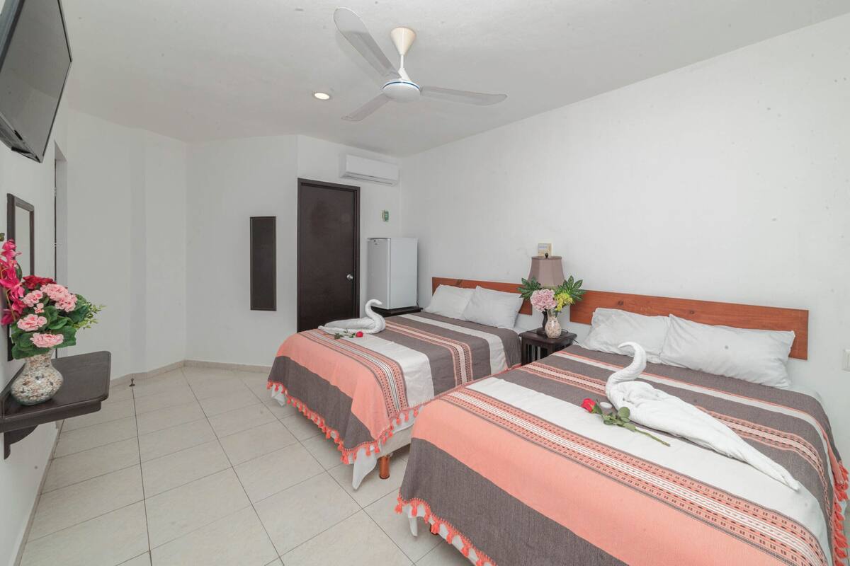 Ein Hotelzimmer mit zwei Betten, einem Deckenventilator, einem Fernseher und einem kleinen Tisch mit Blumen.