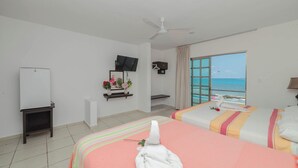 A hotel room with two beds, a TV, a ceiling fan, a window with a view of the sea, and a small desk with a plant. - Hotel La Trigueña (Isla Mujeres)