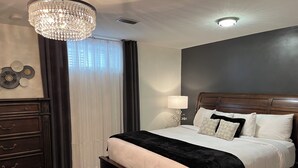 A neatly arranged bedroom with a large bed, a dresser, a chandelier, and a window with curtains.