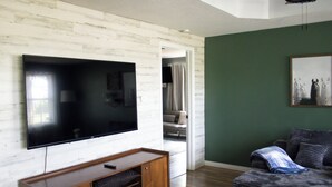 Ein Wohnzimmer mit einem an einer weißen Holzwand montierten Flachbildfernseher, einer Holzunterhaltungseinheit darunter, einer grünen Akzentwand und einem grauen Sofa mit einer Decke.