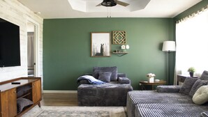 Ein Wohnzimmer mit grüner Wand, einem Flachbildfernseher, einem Sofa, einem Zweisitzer sowie einem Couchtisch und einer Stehlampe.