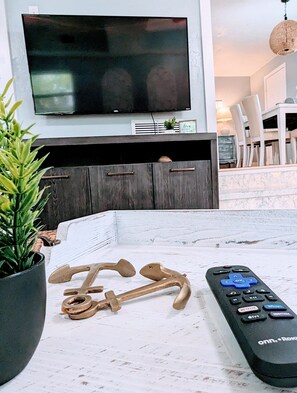 A living room with a TV mounted on the wall, a wooden coffee table, a remote control, and a plant. - SRQ Beach Cottage (Sarasota)