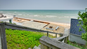 A beach view from a wooden deck with a sandy beach, a wooden fence, and a small building in the background. - 'Summer Place' A Vibrant Rental on Lake Huron (Tawas City)
