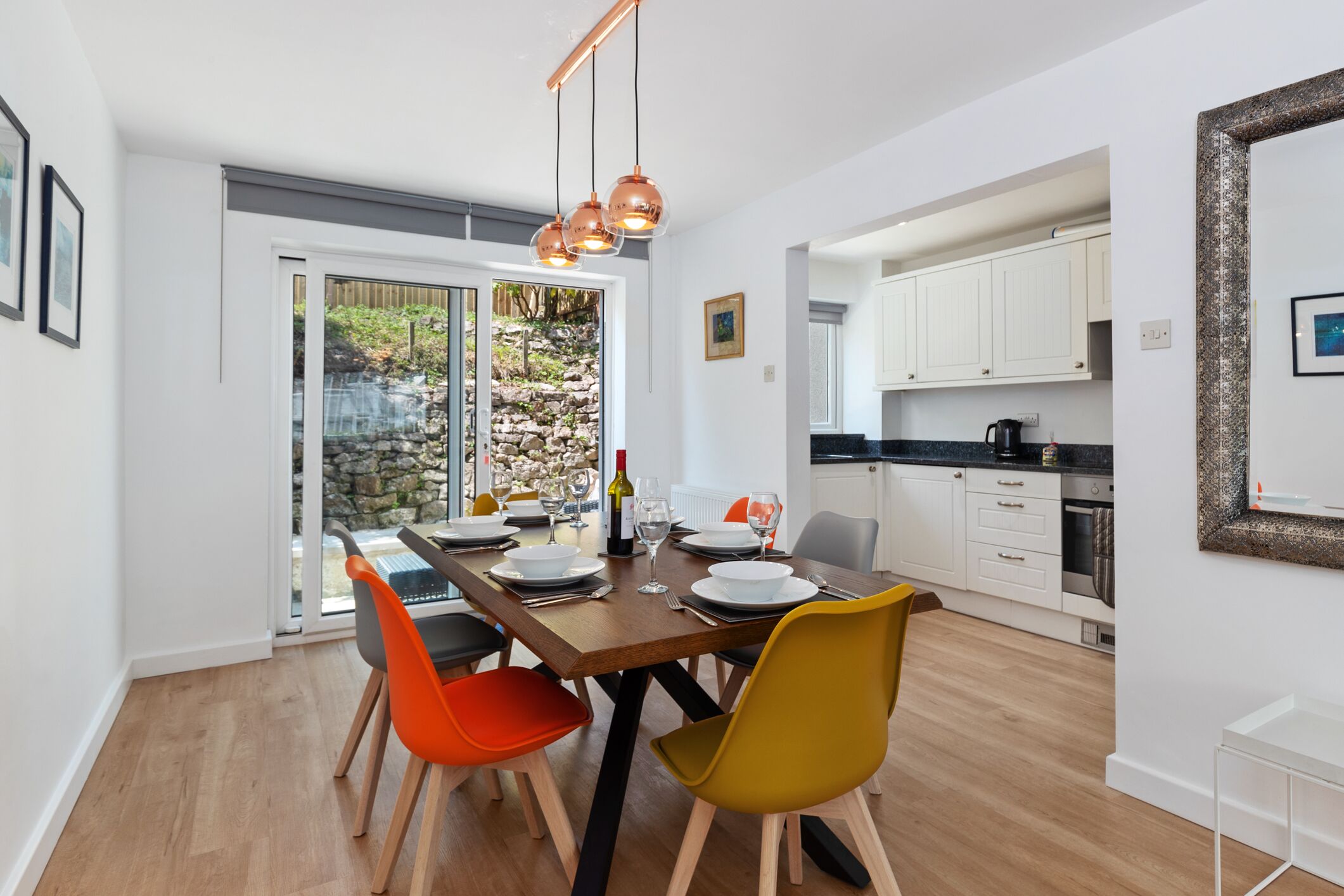 A modern dining area with a wooden table, orange and yellow chairs, and a view of a stone garden through glass doors.