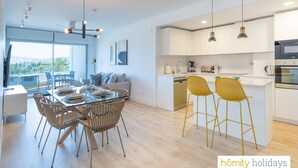 Una cocina moderna con armarios blancos, una isla central con taburetes y una zona de comedor con mesa de vidrio y sillas de mimbre.
