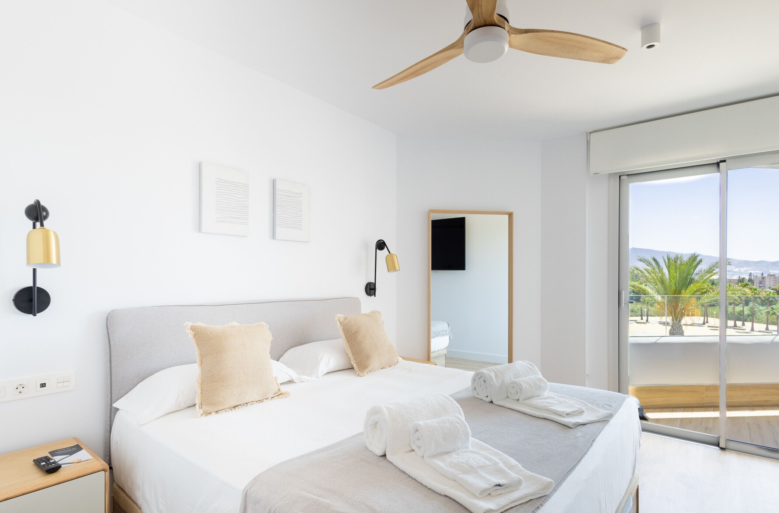 Una habitación de hotel moderna con una cama grande, dos mesitas de noche, un ventilador de techo, un ventanal con vistas y un televisor montado.
