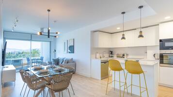 Una cocina moderna con armarios blancos, una isla central con taburetes y una zona de comedor con mesa de vidrio y sillas de mimbre.