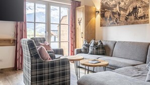Un salon moderne avec un canapé, un fauteuil et deux petites tables. Une grande fenêtre offre une vue sur les montagnes.