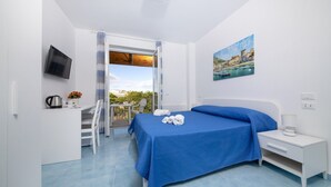A modern bedroom with a large bed, a desk, and a chair. There is a balcony with a view of the sea and buildings. - La Vigna Rooms (Ischia)