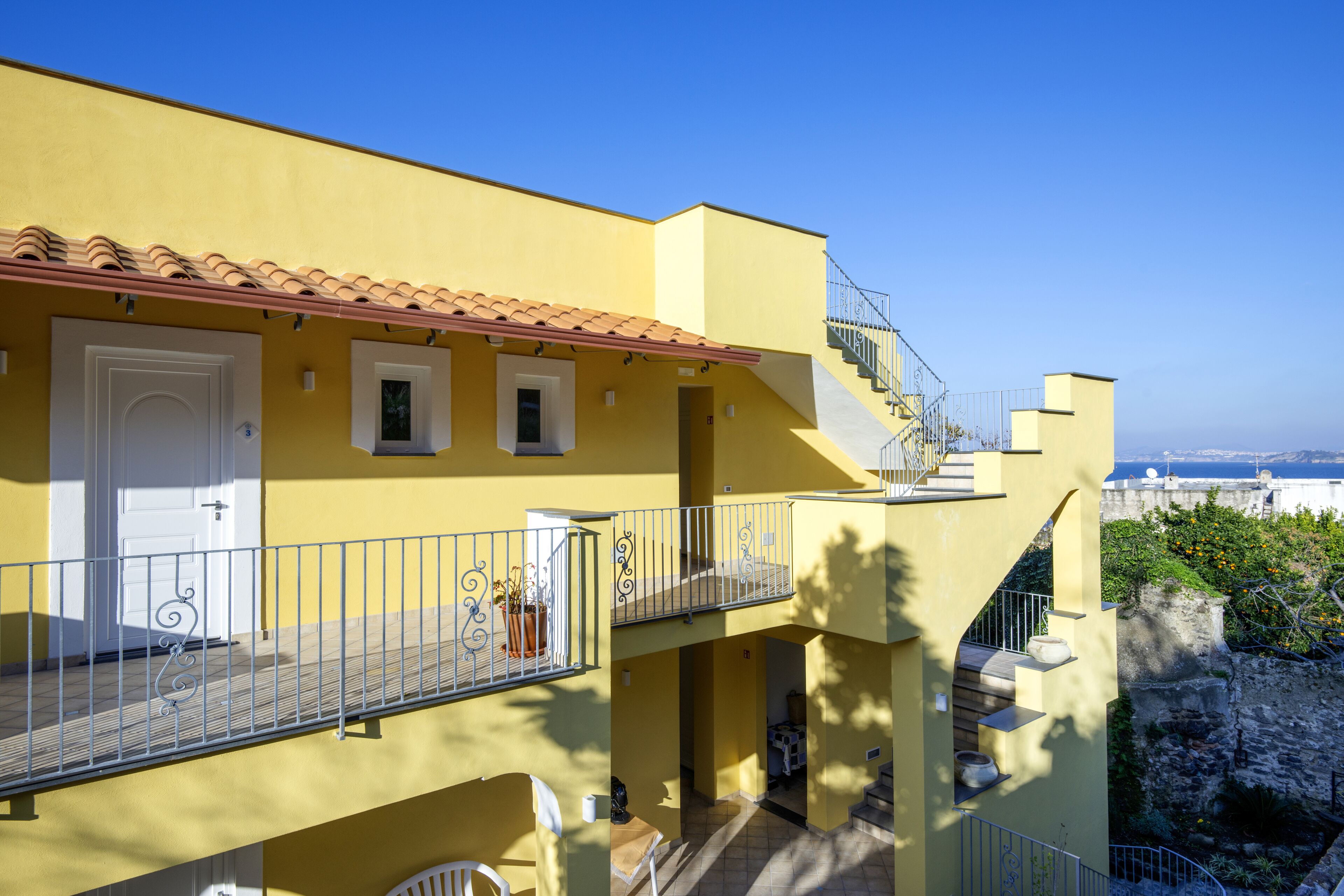 A yellow building with a red-tiled roof, a balcony with metal railings, and a staircase leading to an upper level.