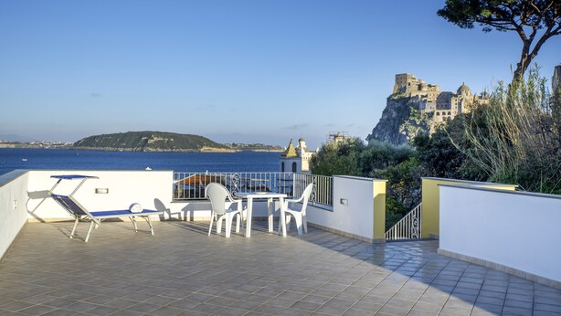 A terrace with a table and chairs, overlooking a coastal area with a castle on a cliff. - La Vigna Rooms (Ischia)