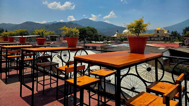 Outdoor seating with tables and chairs, potted plants, and a view of mountains and a yellow building. - Amura Hostel  (Antigua Guatemala)
