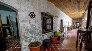 A rustic interior with a wooden ceiling, a worn wall with a ship's wheel decoration, a window with a metal grill, and a tiled floor. - Amura Hostel  (Antigua Guatemala)