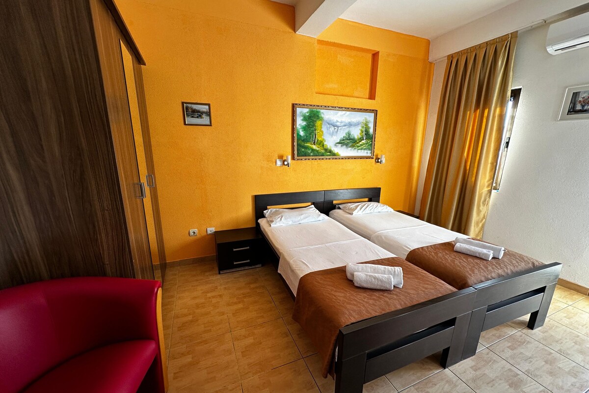 A hotel room with two beds, a red chair, a painting on the wall, and a window with curtains.