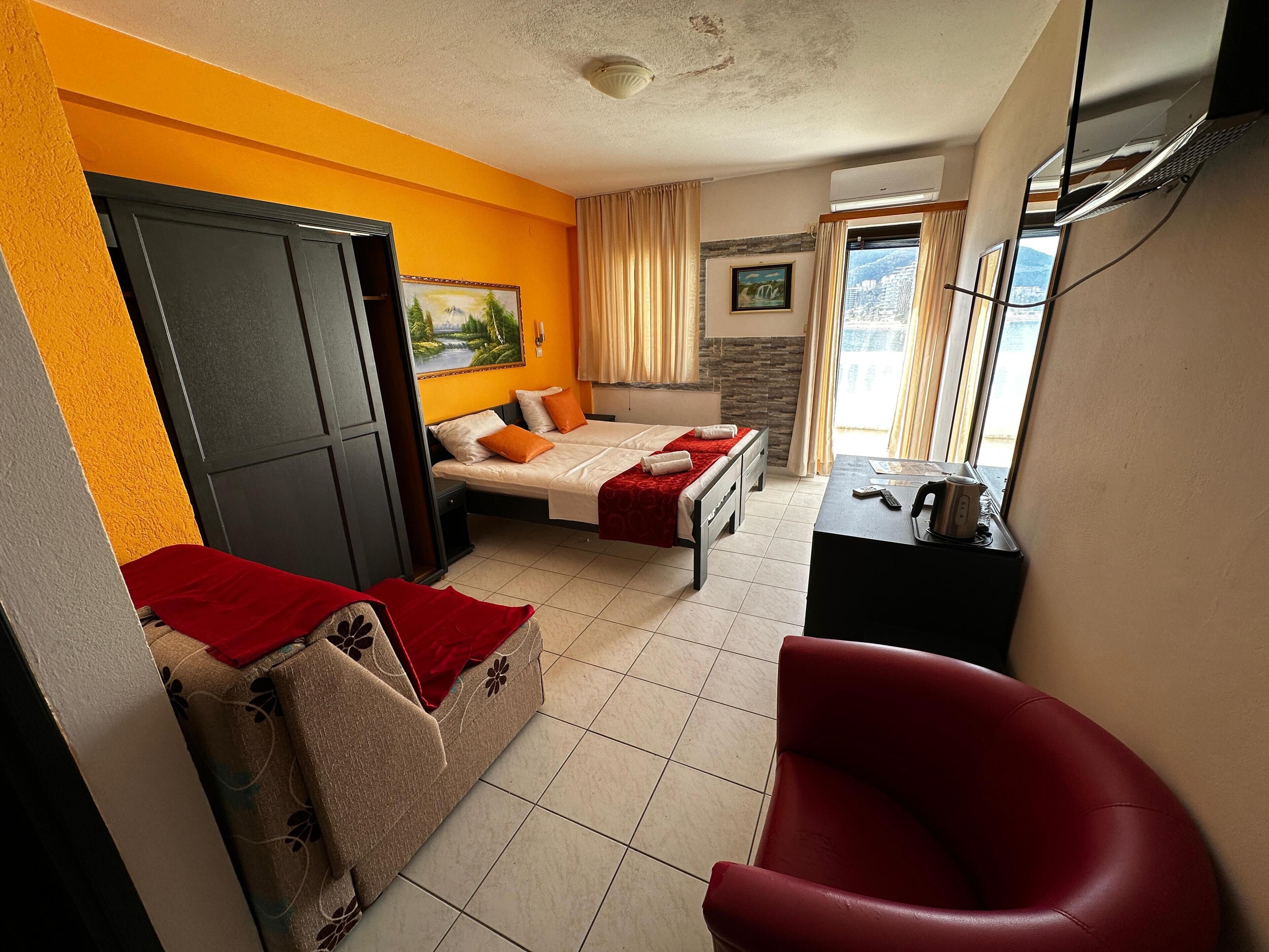 A hotel room with two beds, a sofa, a red chair, a desk with a kettle, and a view of the city through the window.