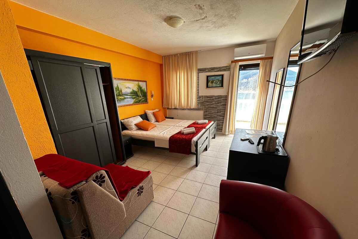 A hotel room with two beds, a sofa, a red chair, a desk with a kettle, and a view of the city through the window.