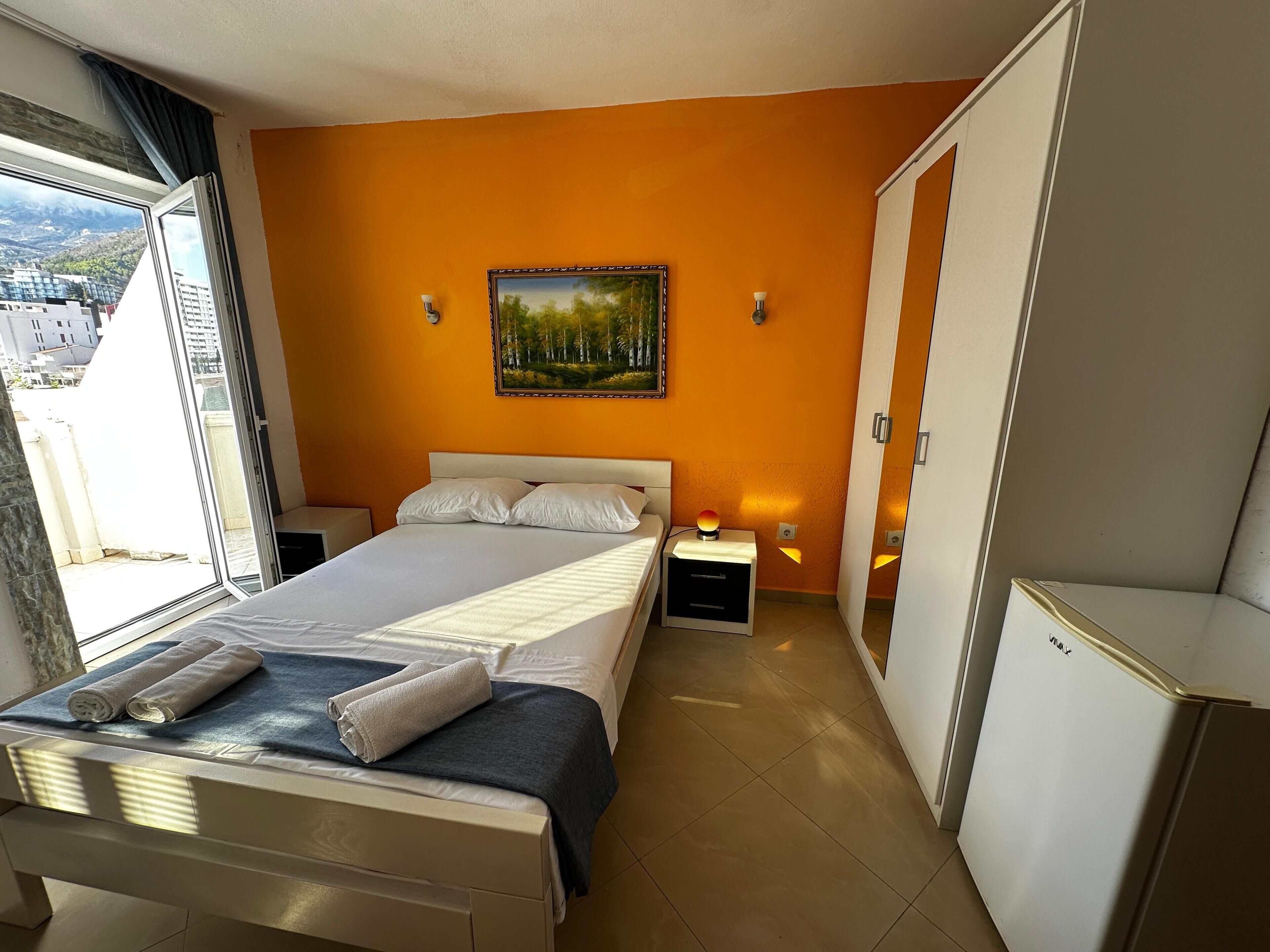 A bedroom with a bed, two bedside tables, a nightstand, a painting on the wall, and a view through the window.