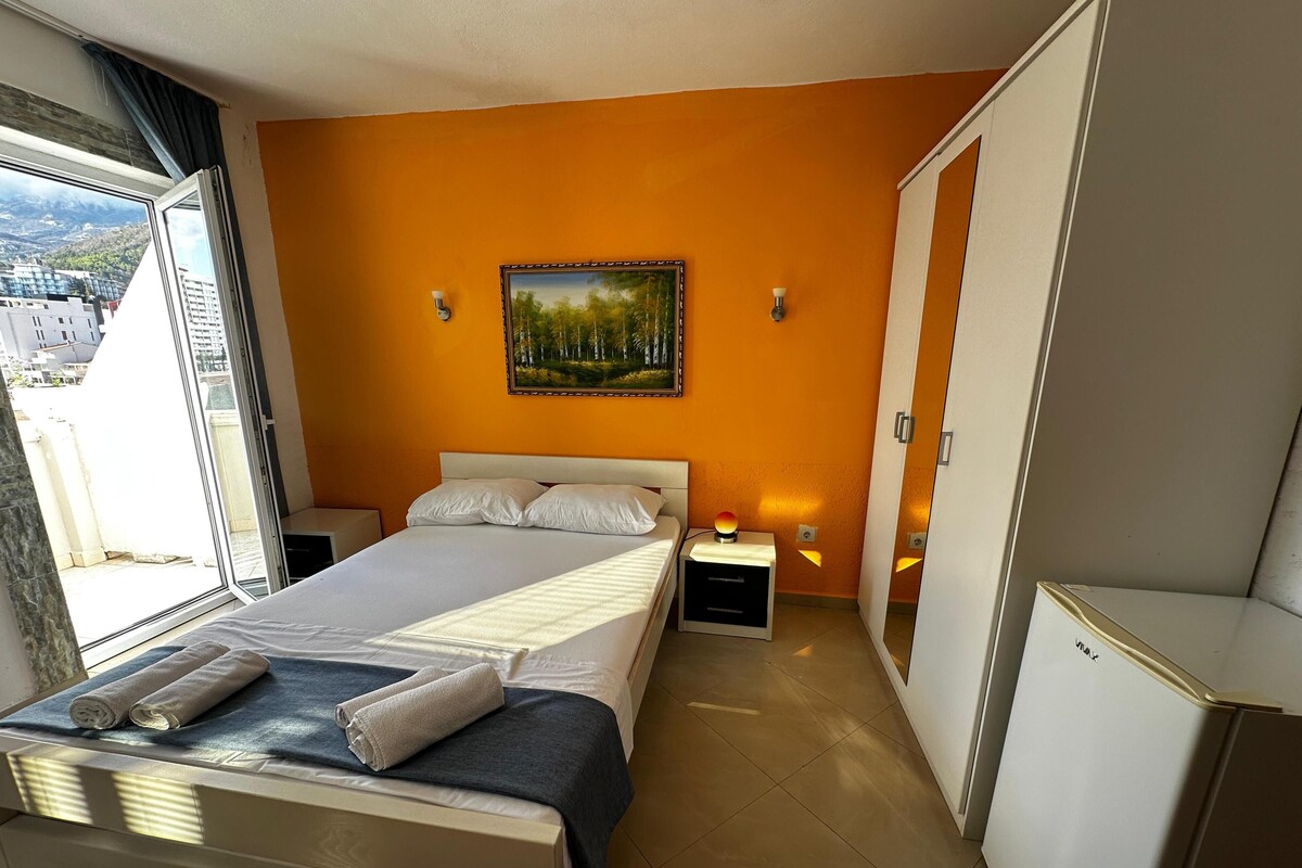 A bedroom with a bed, two bedside tables, a nightstand, a painting on the wall, and a view through the window.