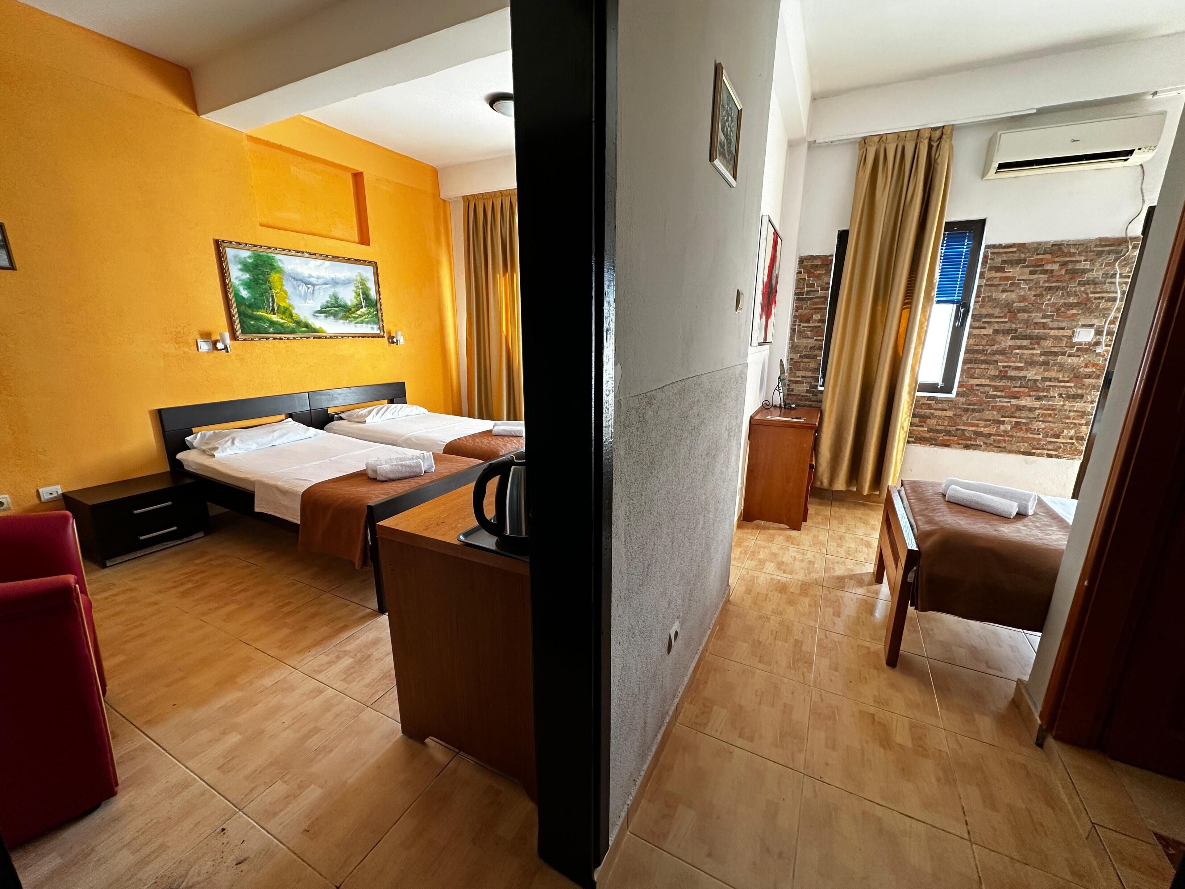 A hotel room with two beds, a desk with a kettle, a red chair, and a view of a brick wall and window with curtains.
