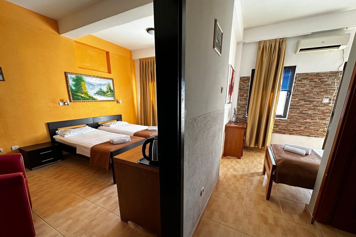 A hotel room with two beds, a desk with a kettle, a red chair, and a view of a brick wall and window with curtains.