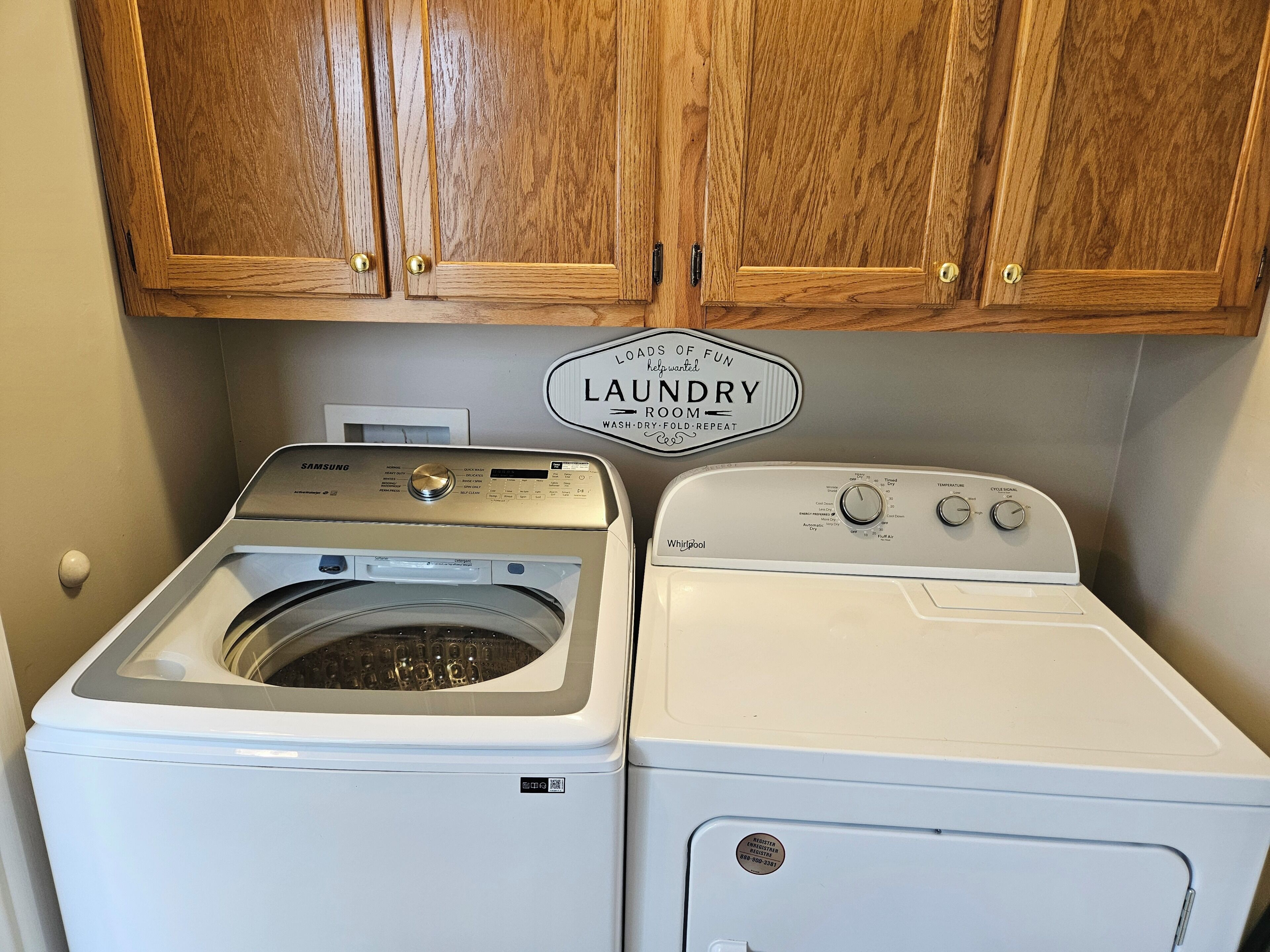 Un lavadero con lavadora Samsung y secadora Whirlpool, armarios de madera arriba y un cartel que indica el propósito de la sala.