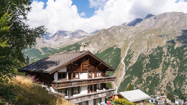 Una casa de estilo chalé en la montaña con balcón, rodeada de exuberante vegetación y montañas.