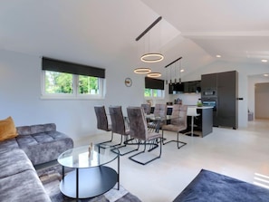 Ein modernes Wohnzimmer mit grauem Sofa, einem Glastisch und einem Essbereich mit Stühlen und Tisch. Es gibt eine Küche mit dunklen Schränken und einer großen Kochinsel.