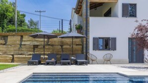 Une piscine avec des chaises longues et des parasols, un mur en pierre et un bâtiment avec des volets.