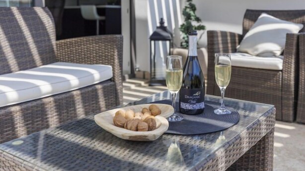 Un canapé en osier avec un coussin, une table en verre avec une bouteille de vin et deux verres, et une assiette de noix.