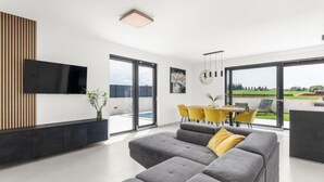 A modern living room with a grey sectional sofa, a flat-screen TV mounted on a wooden panel, and a dining area with yellow chairs and a table. - Villa Valero by Interhome (Novigrad (Istra))