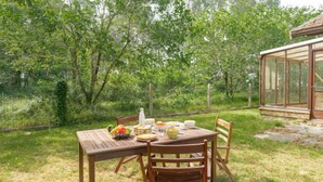 A wooden table set outdoors with a glass enclosure, surrounded by greenery.