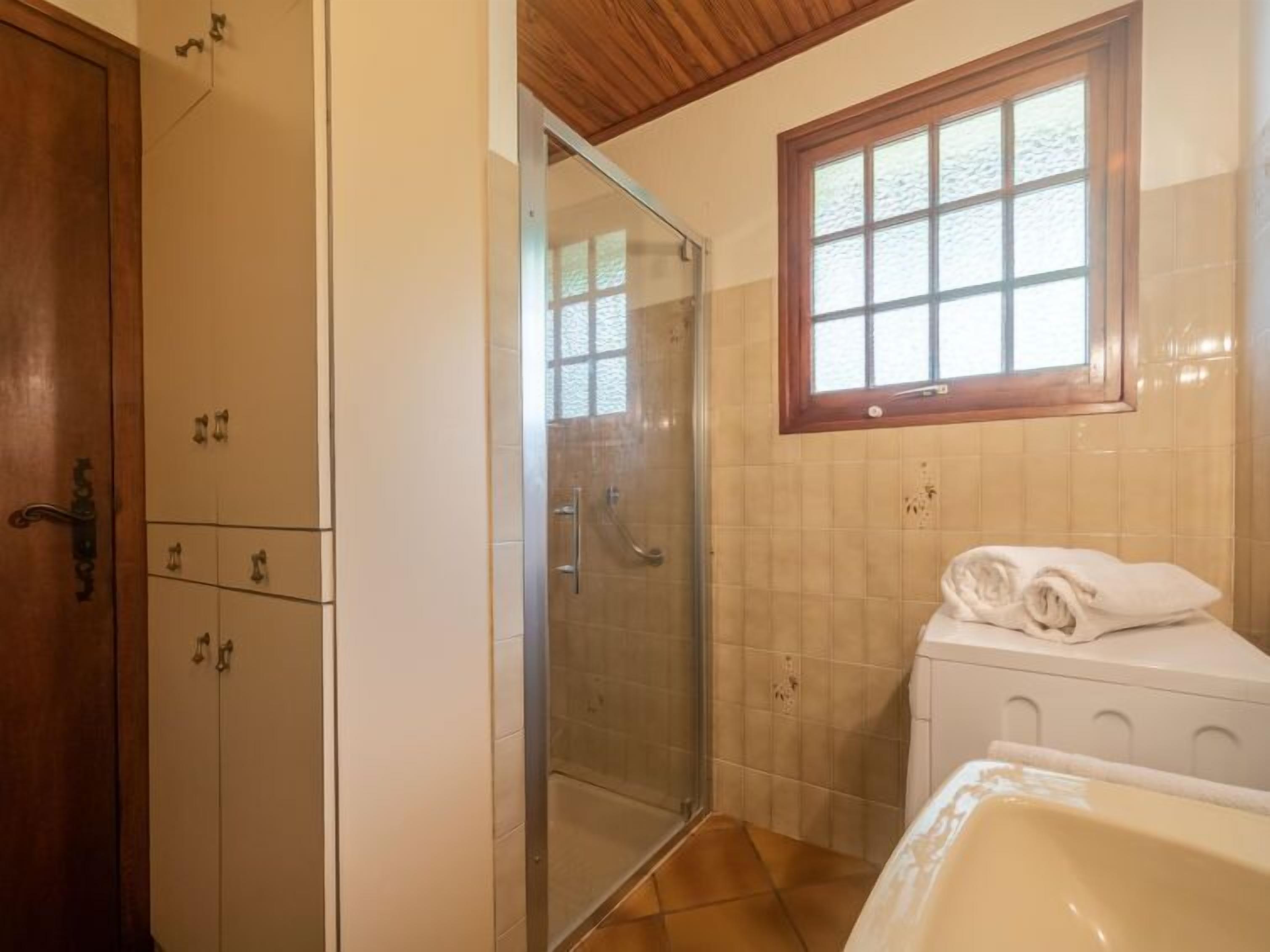 Une salle de bain avec une douche à parois de verre, une porte en bois, une fenêtre avec un cadre en bois et un lavabo blanc sur lequel est posée une serviette.