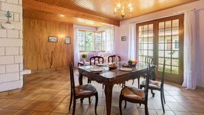 A dining area with a wooden ceiling, a large table, and chairs. There are windows with curtains, a chandelier, and a view of greenery outside.