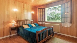 A wooden-paneled bedroom with a bed, a nightstand, a window with a view of greenery, and a radiator.
